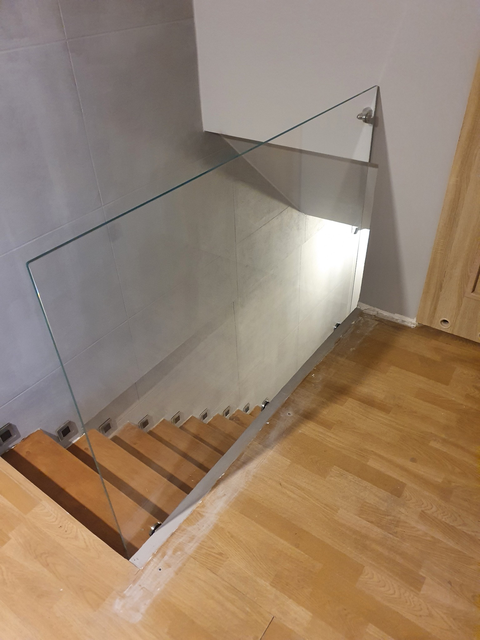 Szklana balustrada zamocowana na drewnianych schodach, widok z góry, minimalistyczny design, szare płytki na ścianie w tle, jasna drewniana podłoga.