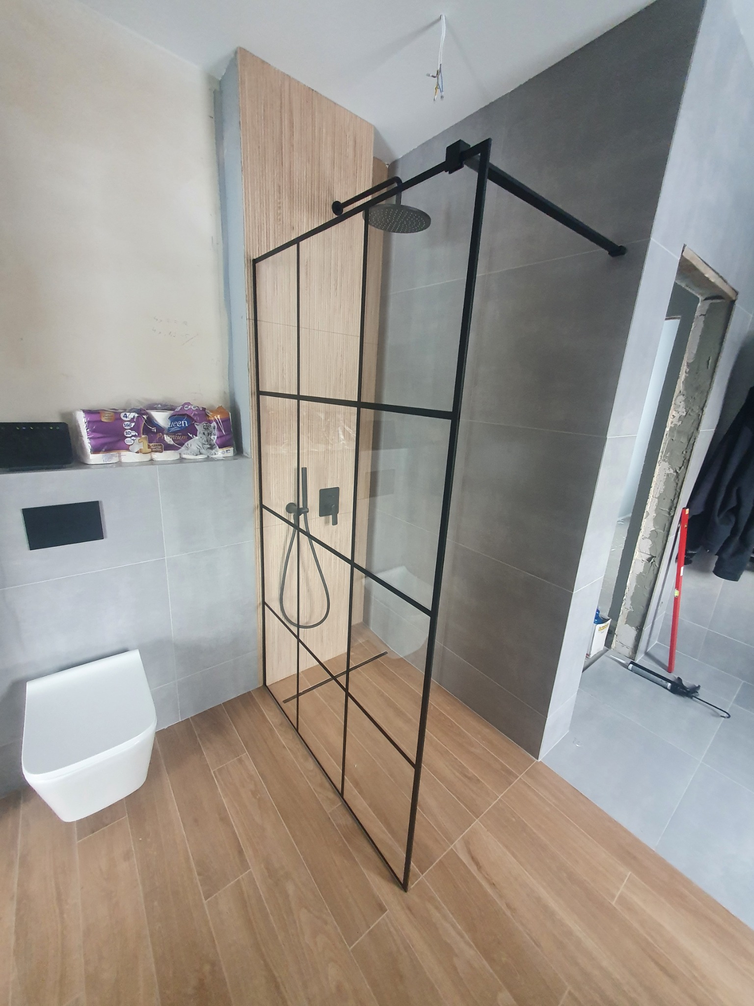 Kabina prysznicowa w stylu loft z czarnymi profilami i drewnianym panelem ściennym, zamontowana w łazience z szarą glazurą i drewnopodobną podłogą, widoczny fragment toalety i zapas papieru...