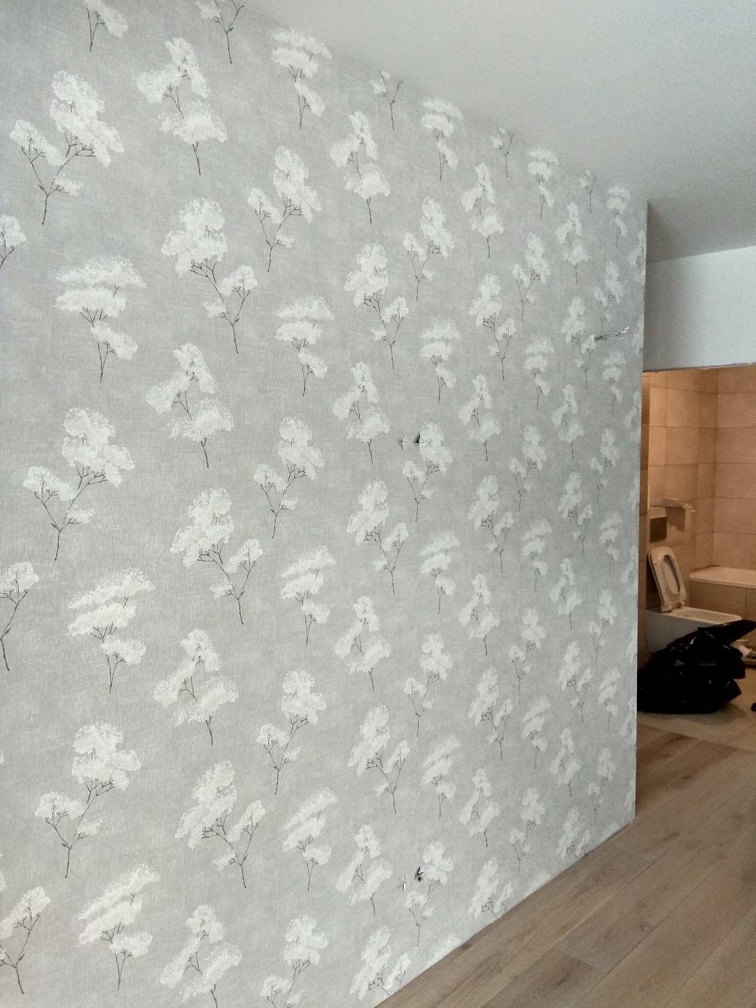 Ściana pokryta jasnoszarą tapetą w drobne białe kwiatki, widoczne niedoskonałości w postaci dziurek i nierówności, fragment podłogi z paneli oraz łazienka w tle.