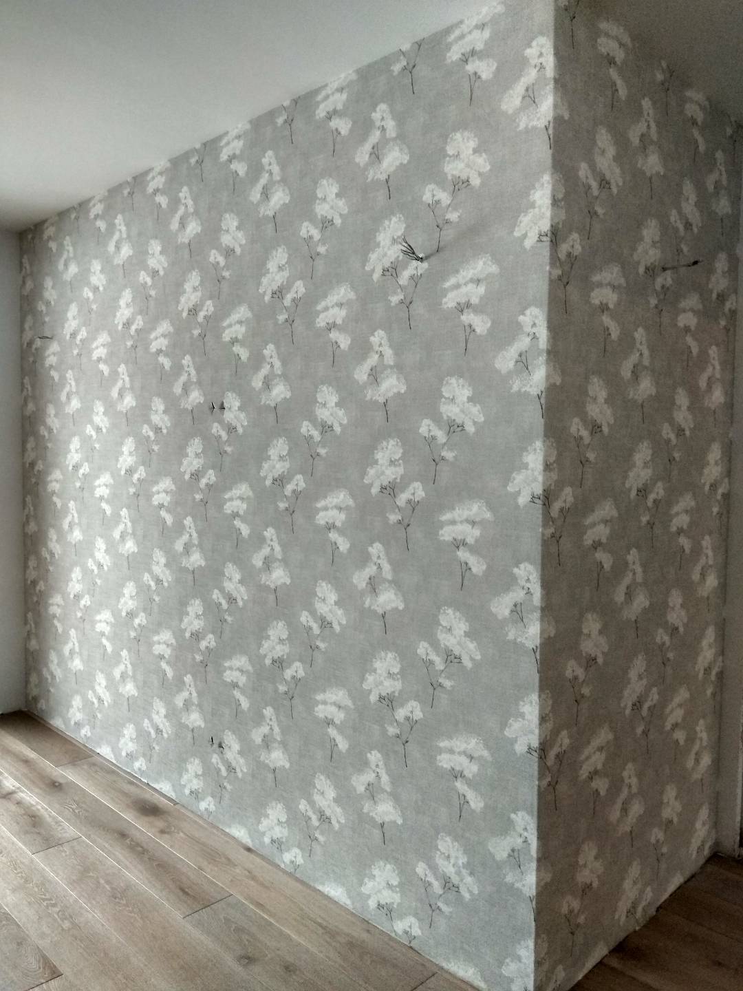 Wykończona ściana w narożniku pokoju, pokryta szarą tapetą w białe kwiatowe wzory, z widocznym fragmentem drewnianej podłogi.