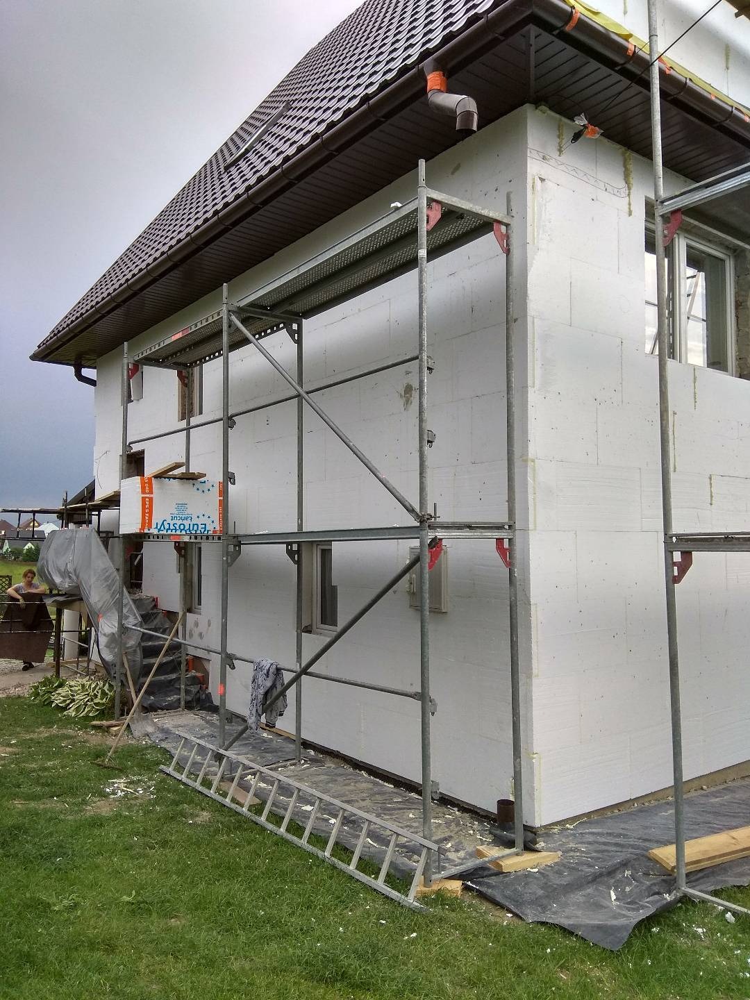 Dom jednorodzinny w trakcie ocieplania styropianem, widoczne rusztowania, fragment okna i brązowy dach z rynnami, na trawie leżą resztki styropianu, pochmurne niebo.