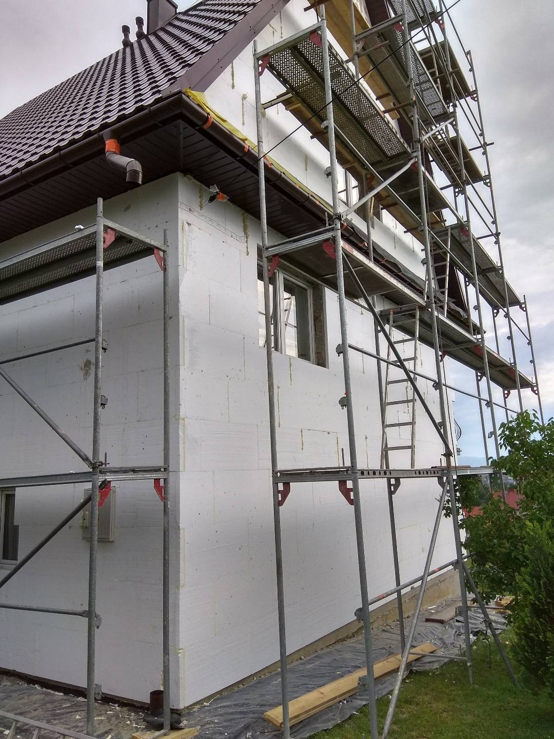 Dom jednorodzinny w trakcie ocieplania elewacji, widoczne rusztowania i warstwa styropianu, brązowy dach z rynnami, częściowo zakryte okno, zabezpieczona folią podstawa budynku.