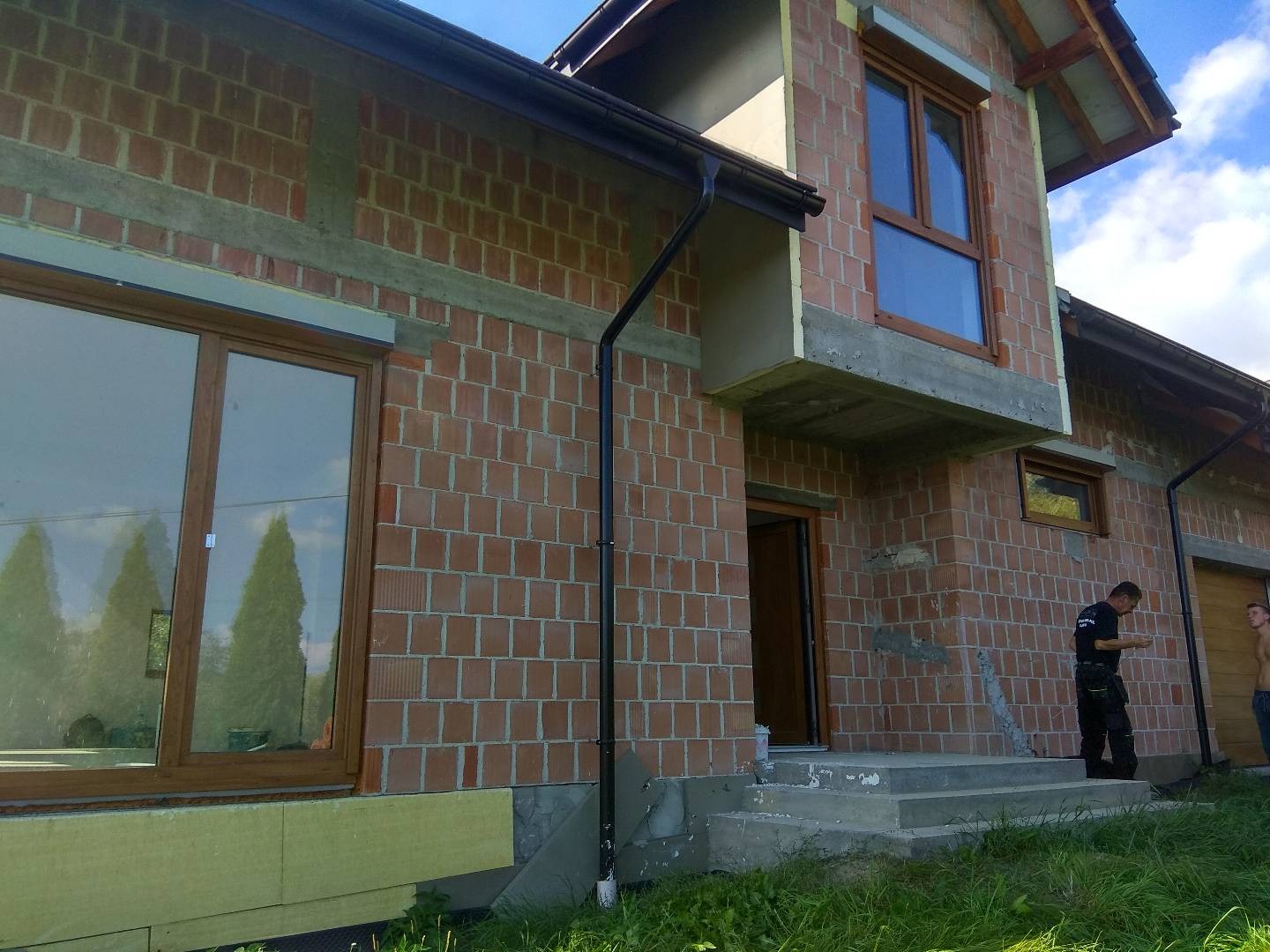 Surowy budynek w trakcie budowy z czerwonej cegły, z oknami z brązowymi ramami, częściowo ocieplony, z widocznymi pracownikami na zewnątrz.