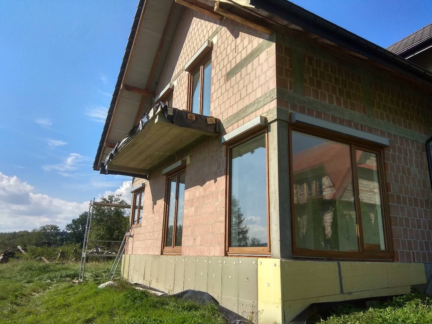 Dom jednorodzinny w trakcie prac elewacyjnych: widoczne ocieplenie fundamentów żółtym styropianem, fragment rusztowania oraz niezakończone obróbki okien.