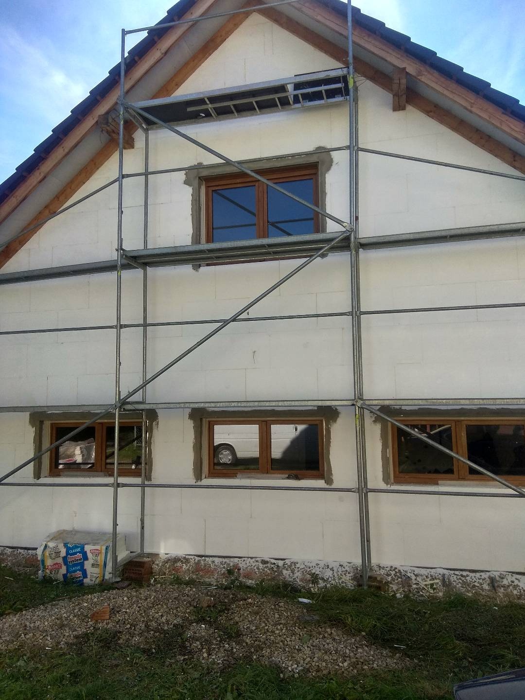 Dom w trakcie ocieplania: ściana zewnętrzna z białych płyt, częściowo osadzona w zaprawie wokół brązowych okien, rusztowanie metalowe, worki z materiałem budowlanym u podstawy, trawa i kamienie...