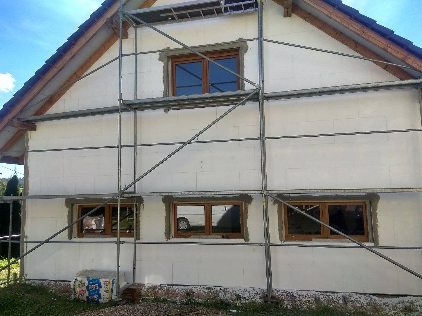 Dom w trakcie ocieplania z widocznymi rusztowaniami, styropianem i workiem zaprawy murarskiej u podstawy ściany, okna z drewnianymi ramami częściowo osadzone.