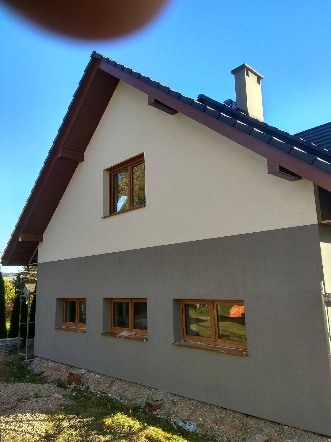 Dwukolorowa elewacja domu jednorodzinnego z drewnianymi oknami, widok z boku na tle błękitnego nieba i fragmentu ogrodu, widoczne niedokończone prace wykończeniowe wokół budynku.