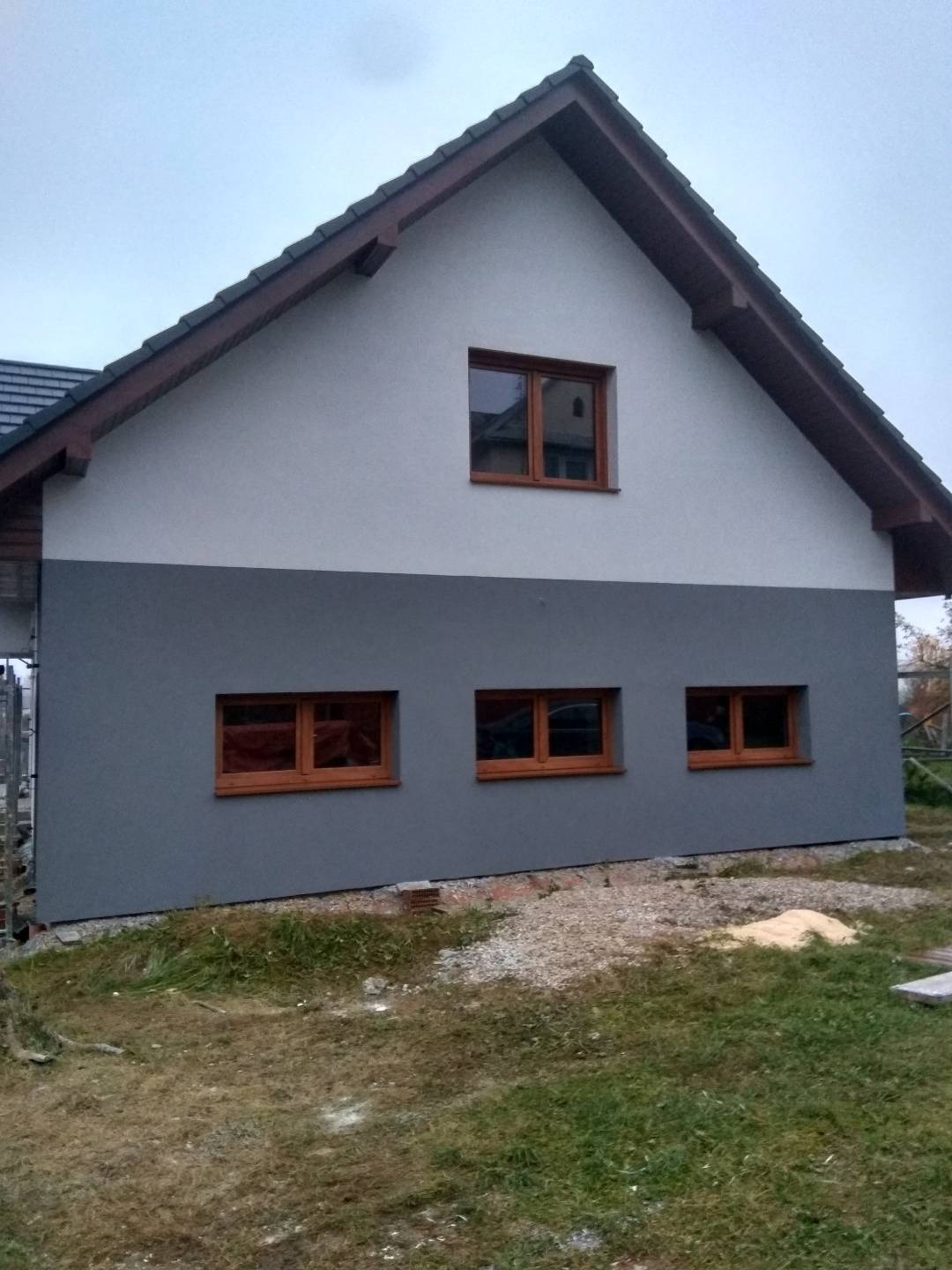 Dwukolorowa elewacja domu jednorodzinnego z trzema brązowymi oknami na dole i jednym u góry, widok z boku, trawa i kamienie u podstawy.