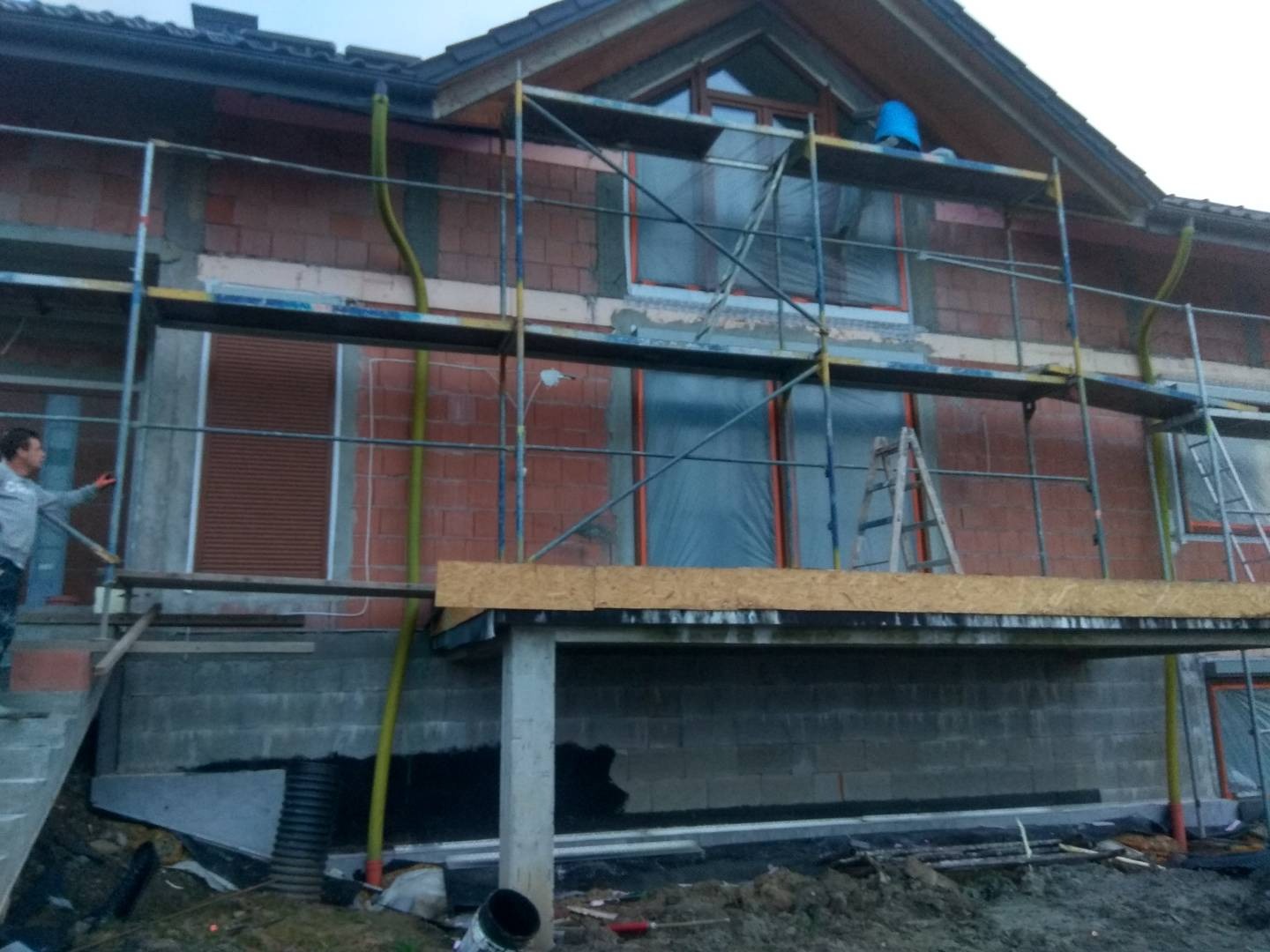 Dom w trakcie ocieplania elewacji, widoczne rusztowania, częściowo ocieplona ściana z cegły, okna zabezpieczone folią, pracownik na schodach.