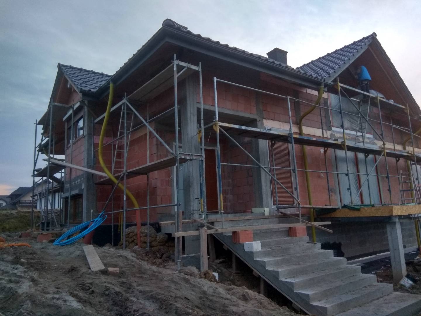 Dom w trakcie budowy, widoczne rusztowania, niezabezpieczone okna, ściany z czerwonej cegły, betonowe schody wejściowe i pracownik w niebieskiej kurtce na rusztowaniu