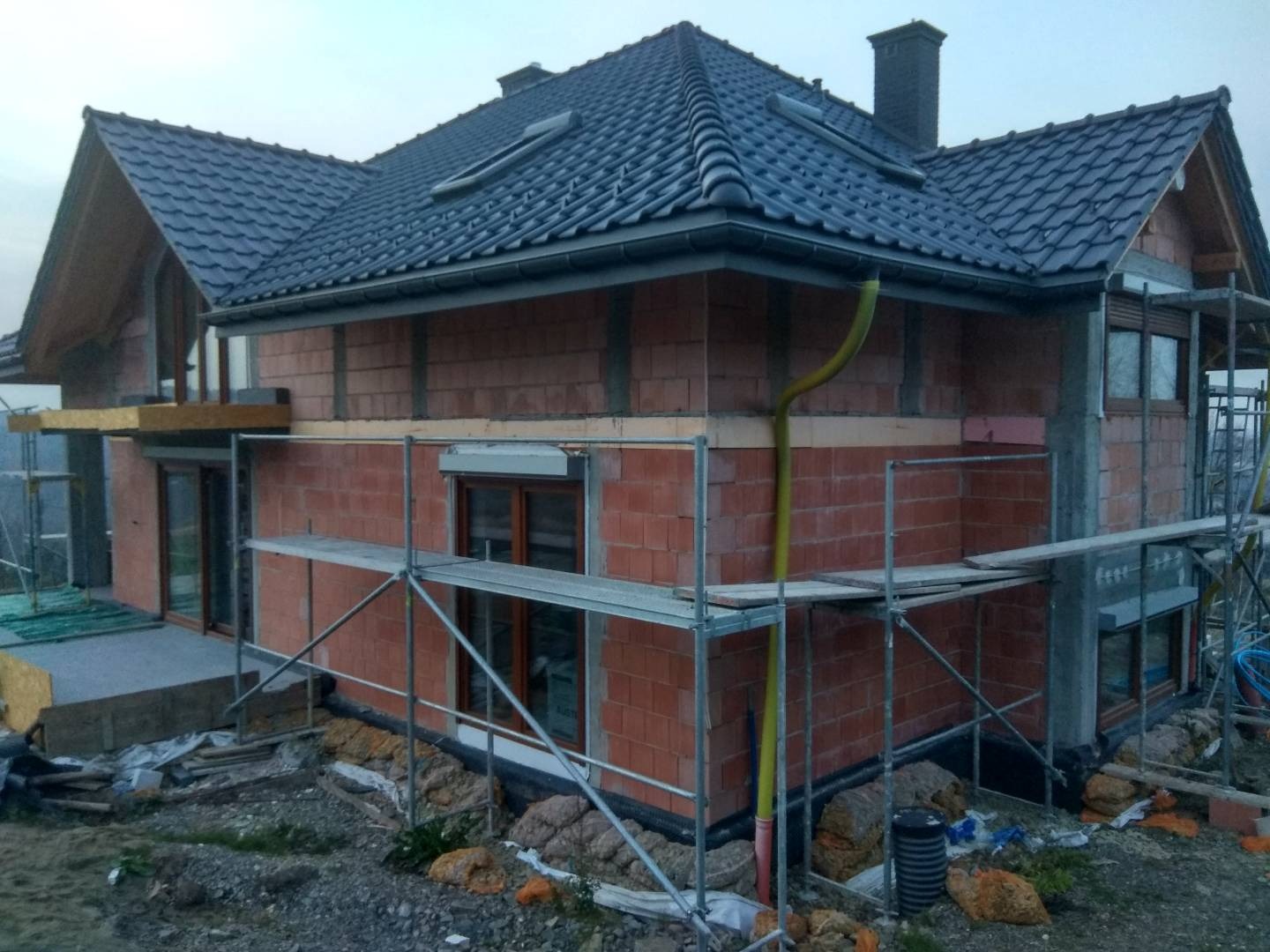 Dom w trakcie budowy: widoczne niezabezpieczone ściany z cegły, rusztowania, okna z roletami zewnętrznymi, dachówka na dachu i rynna spustowa, teren wokół domu niezagospodarowany.