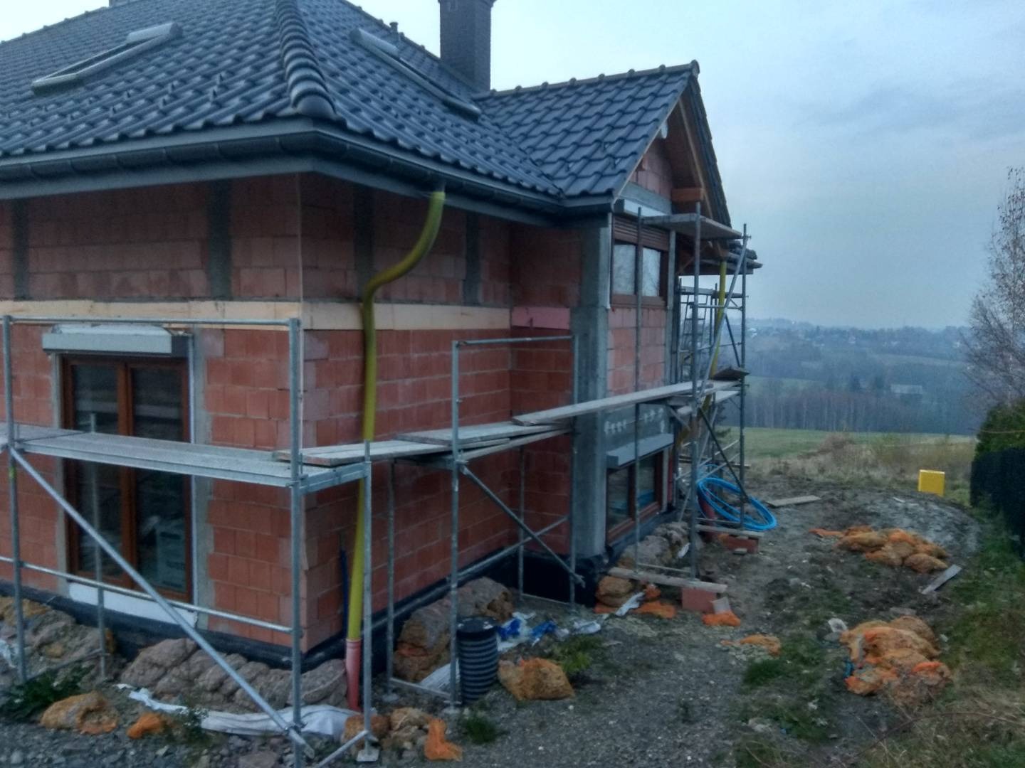 Niedokończony budynek z czerwonej cegły z rusztowaniem, widoczne ocieplenie i rynna w kolorze żółtym, w tle pagórkowaty krajobraz.