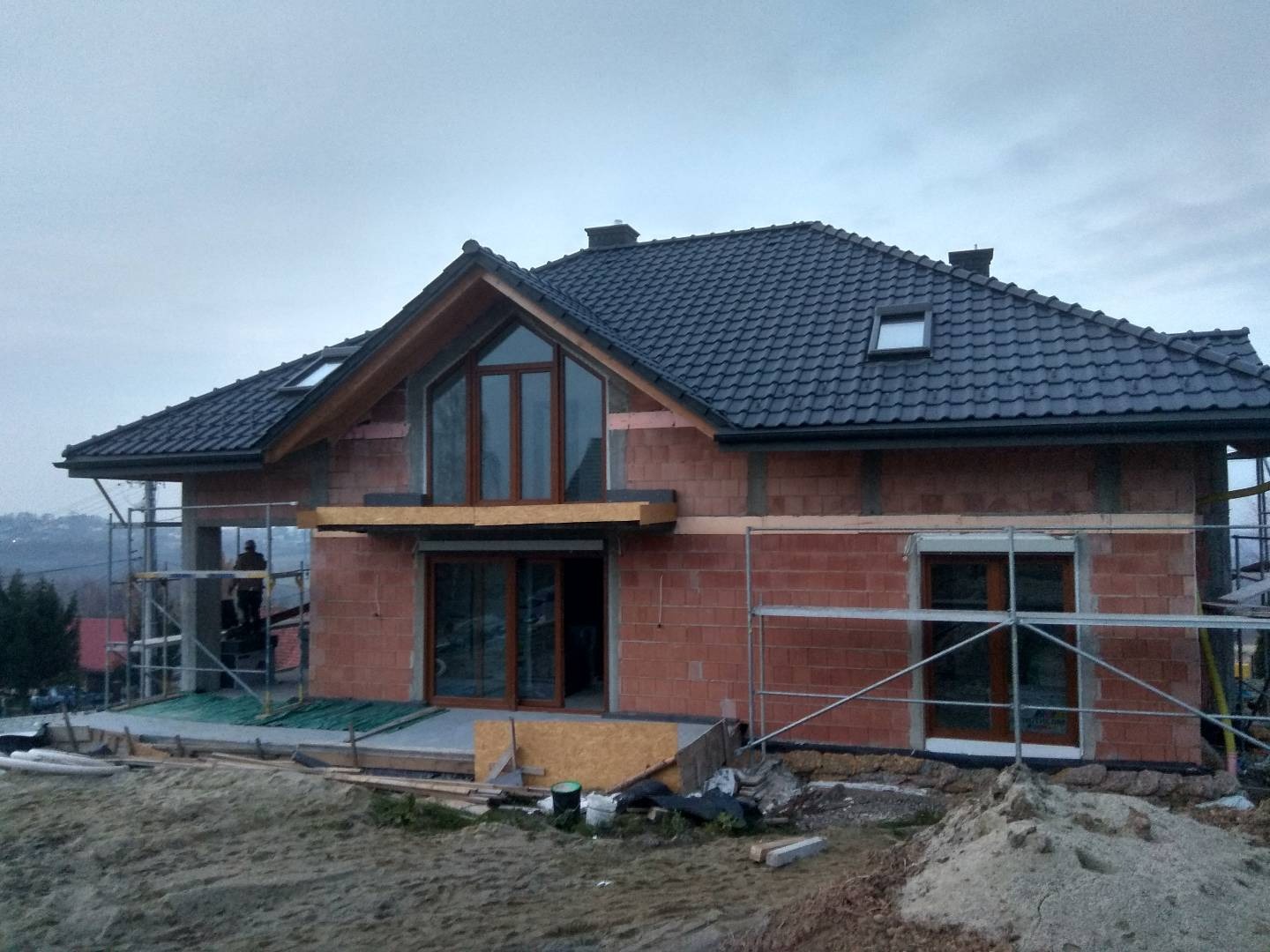 Dom w trakcie budowy: ściany z czerwonej cegły, czarny dach z dachówką, okna z brązowymi ramami, rusztowania metalowe, widoczne ocieplenie na górze ściany szczytowej, teren wokół domu jeszcze...