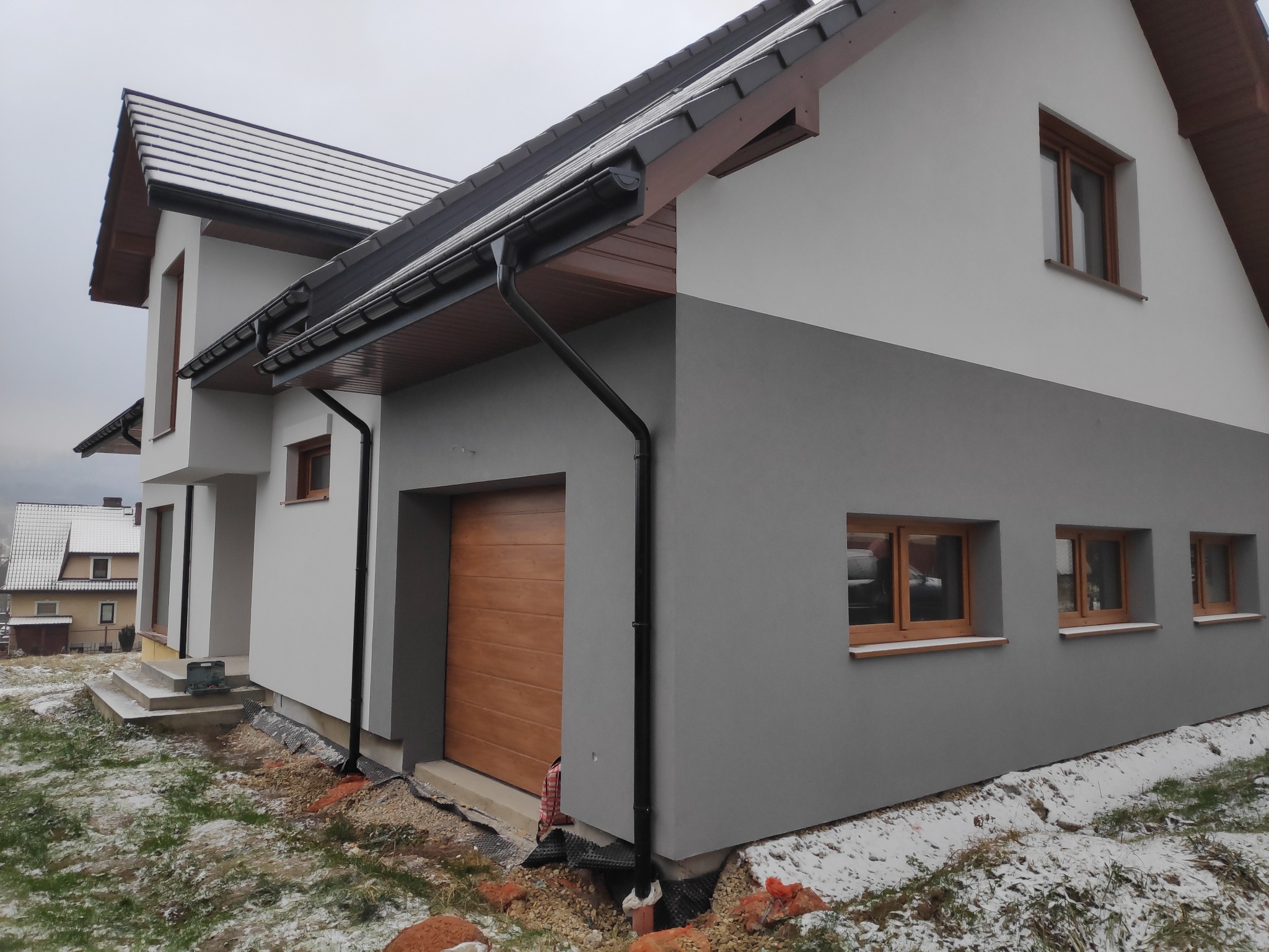 Dwukolorowa elewacja domu jednorodzinnego z brązową stolarką okienną i drewnianą bramą garażową, widoczna w zimowej scenerii z fragmentami śniegu na trawie.