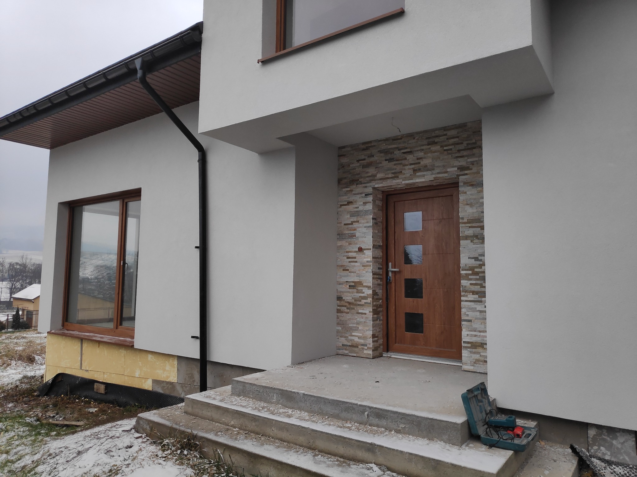 Wykończona elewacja domu jednorodzinnego w Chabówce z ociepleniem i dekoracyjnym kamieniem przy wejściu, widoczne schody wejściowe i skrzynka z narzędziami.