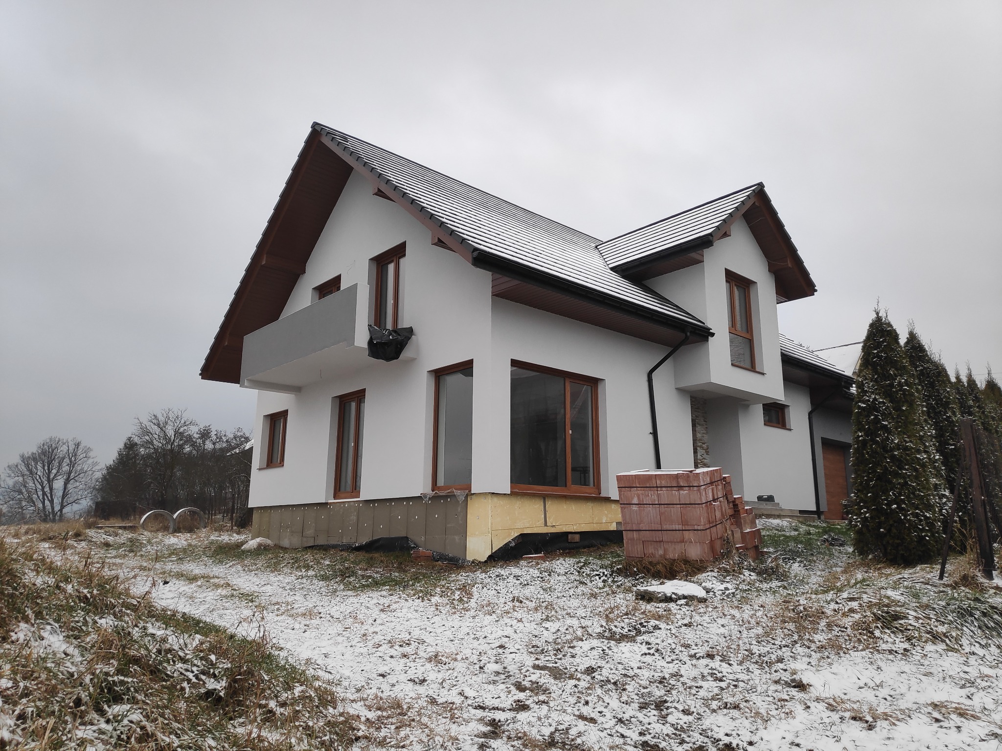 Nowoczesny, dwukondygnacyjny dom z białymi ścianami i brązowymi ramami okiennymi w trakcie ocieplania, widoczne ocieplenie fundamentów i stos cegieł na zewnątrz w zimowej scenerii.