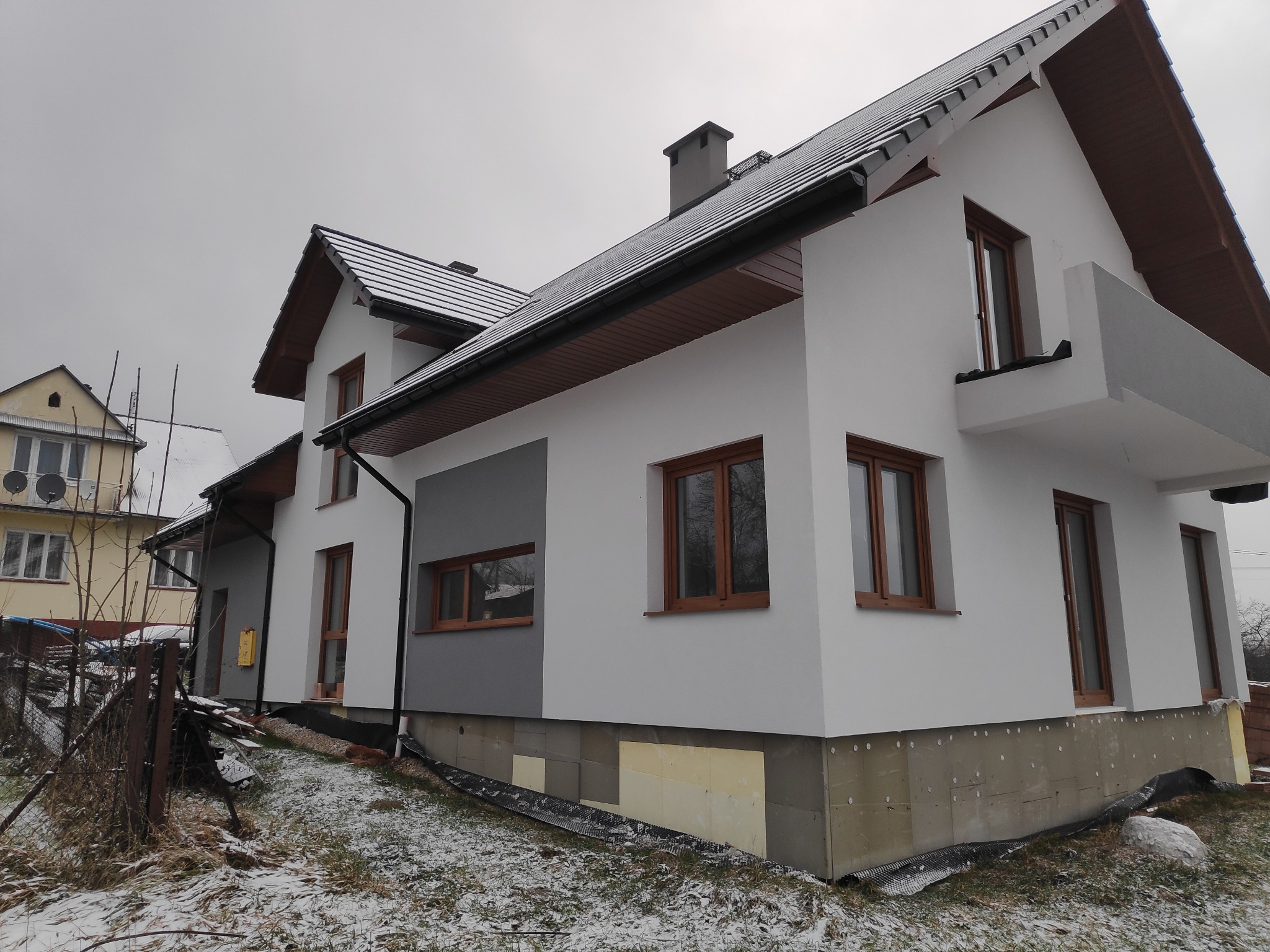 Nowoczesny dom jednorodzinny z ocieploną elewacją, widoczne fragmenty ocieplenia fundamentów, szara kolorystyka fasady z brązowymi ramami okiennymi, pochmurne niebo.