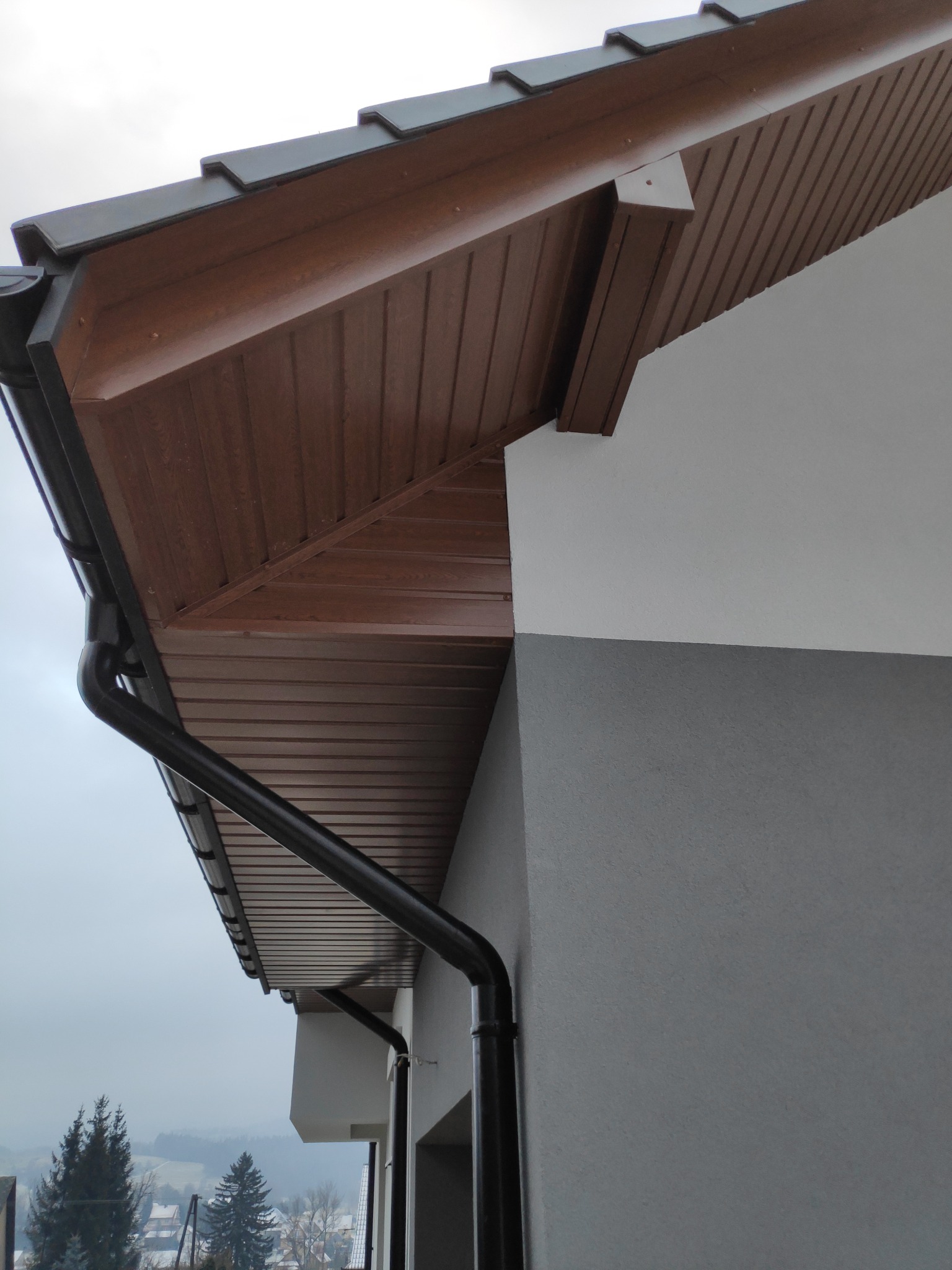 Fragment elewacji budynku z widocznym ociepleniem podbitki dachowej z brązowego panelu PCV, orynnowaniem w kolorze czarnym oraz dwukolorową szarą elewacją.