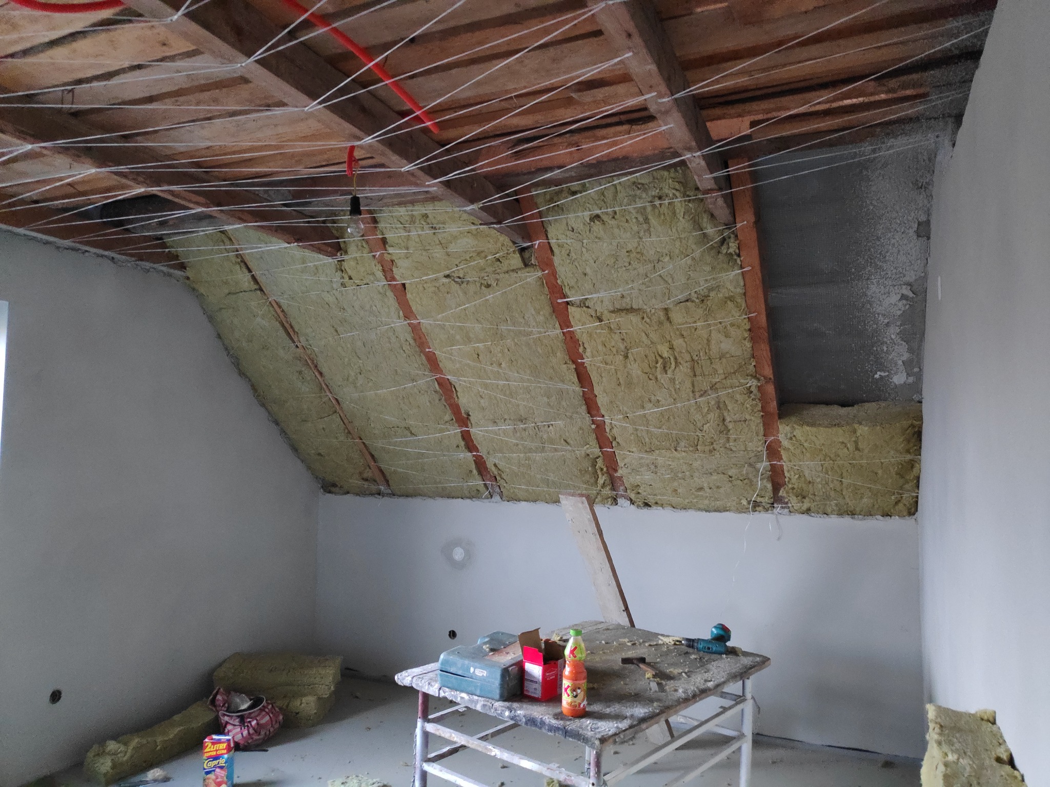 Ocieplenie poddasza wełną mineralną, widoczne drewniane belki, sznurki mocujące, częściowo otynkowana ściana i narzędzia na stole warsztatowym.