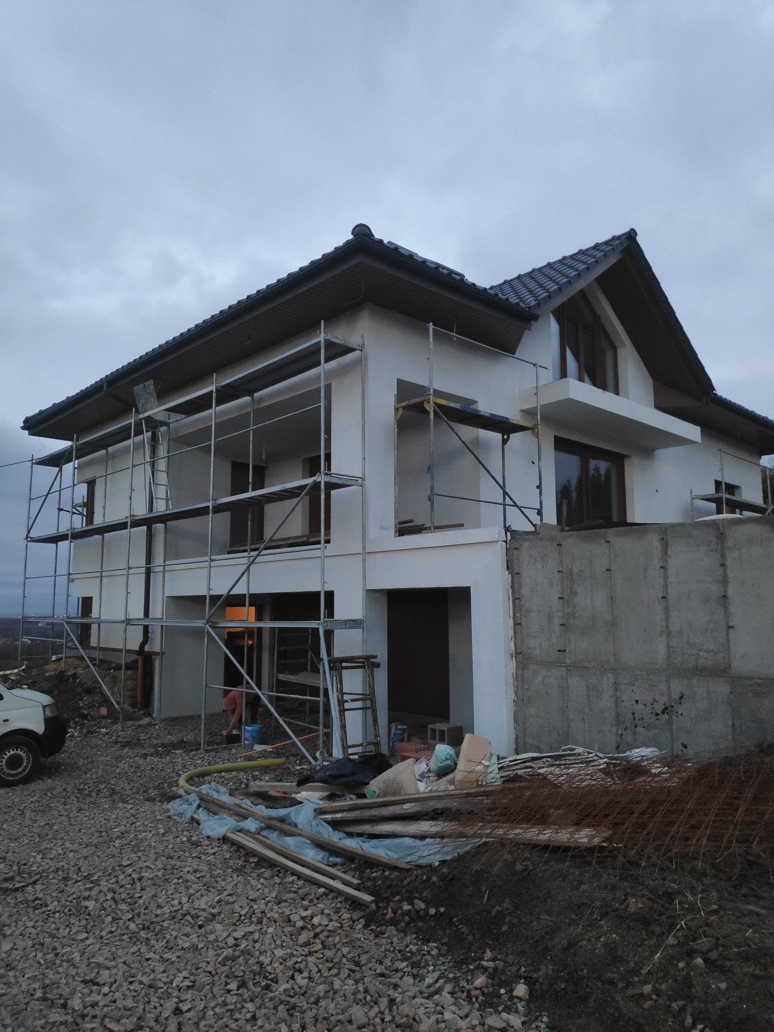 Dom jednorodzinny w trakcie prac wykończeniowych, z widocznym rusztowaniem, częściowo otynkowaną elewacją, niezabudowaną piwnicą i materiałami budowlanymi na posesji