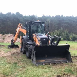 Pomarańczowo-czarna koparko-ładowarka Case 580SL z dużą łyżką z przodu i mniejszą z tyłu, stojąca na trawiastym terenie z widoczną pryzmą ziemi w tle, na obrzeżach lasu.