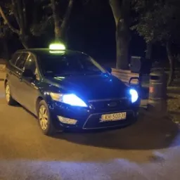 Ciemnozielony samochód kombi z taksometrem na dachu, zaparkowany nocą przy chodniku w parku, widoczna tablica rejestracyjna z Krakowa.