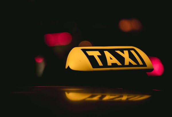 Żółty znak TAXI na dachu samochodu, oświetlony w nocy, z widocznym odbiciem na karoserii i rozmazanymi światłami w tle.
