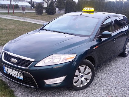 Ciemnozielony Ford Mondeo kombi z żółtym szyldem TAXI na dachu, zaparkowany na żwirowym podjeździe przed domem.