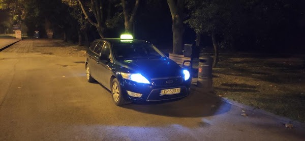 Ciemnozielony samochód kombi z taksometrem na dachu, zaparkowany nocą przy chodniku w parku, widoczna tablica rejestracyjna z Krakowa.