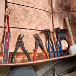 Miedziane panele dachowe w trakcie montażu, narzędzia dekarskie: nożyce do blachy z zielonymi i pomarańczowymi uchwytami, młotek dekarski, miarka Eurovent, widoczne ślady obróbki blachy.