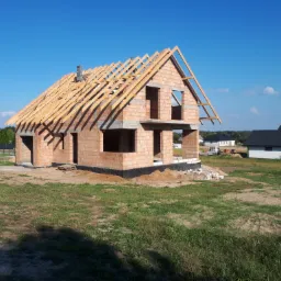 Niedokończony, dwukondygnacyjny budynek z czerwonej cegły z drewnianą konstrukcją dachu, w otoczeniu trawnika i innych budynków w tle, w słoneczny dzień.