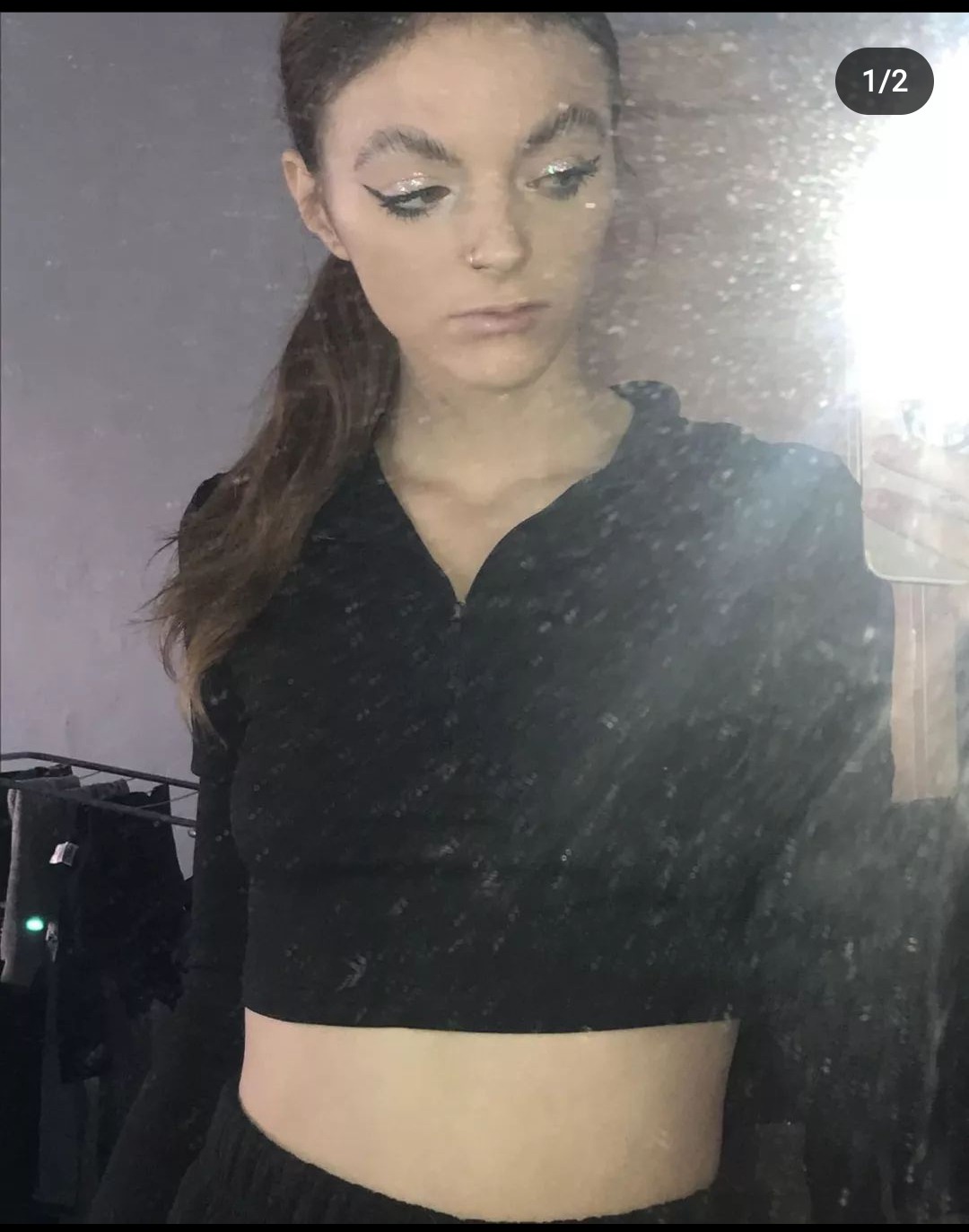 Portret kobiety z brokatowym makijażem oczu i włosami związanymi w kucyk, pozującej w czarnym topie na tle lustra.