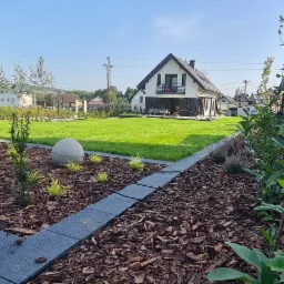 Nowoczesny ogród z równo przystrzyżonym trawnikiem, rabatami wyłożonymi korą, młodymi drzewkami iglastymi i dekoracyjną kulą betonową na tle domu z balkonem.