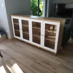 Nowoczesna przeszklona komoda z drewnianymi półkami i białymi ramami, ustawiona na niskich nóżkach w salonie z drewnianą podłogą.