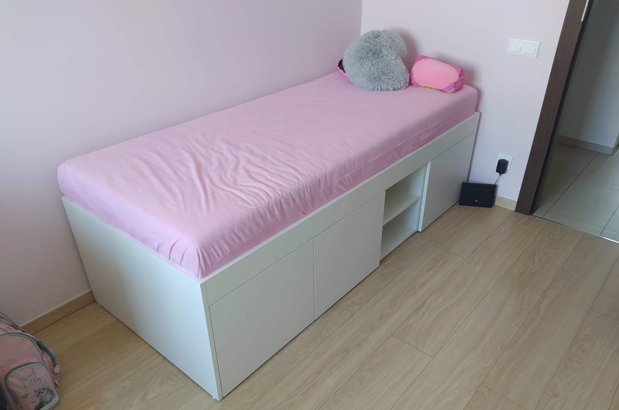 Łóżko jednoosobowe z białym korpusem i różowym materacem, wbudowane szafki z półkami, z widoczną poduszką w kształcie serca i szarą poduszką w tle, na drewnianej podłodze.