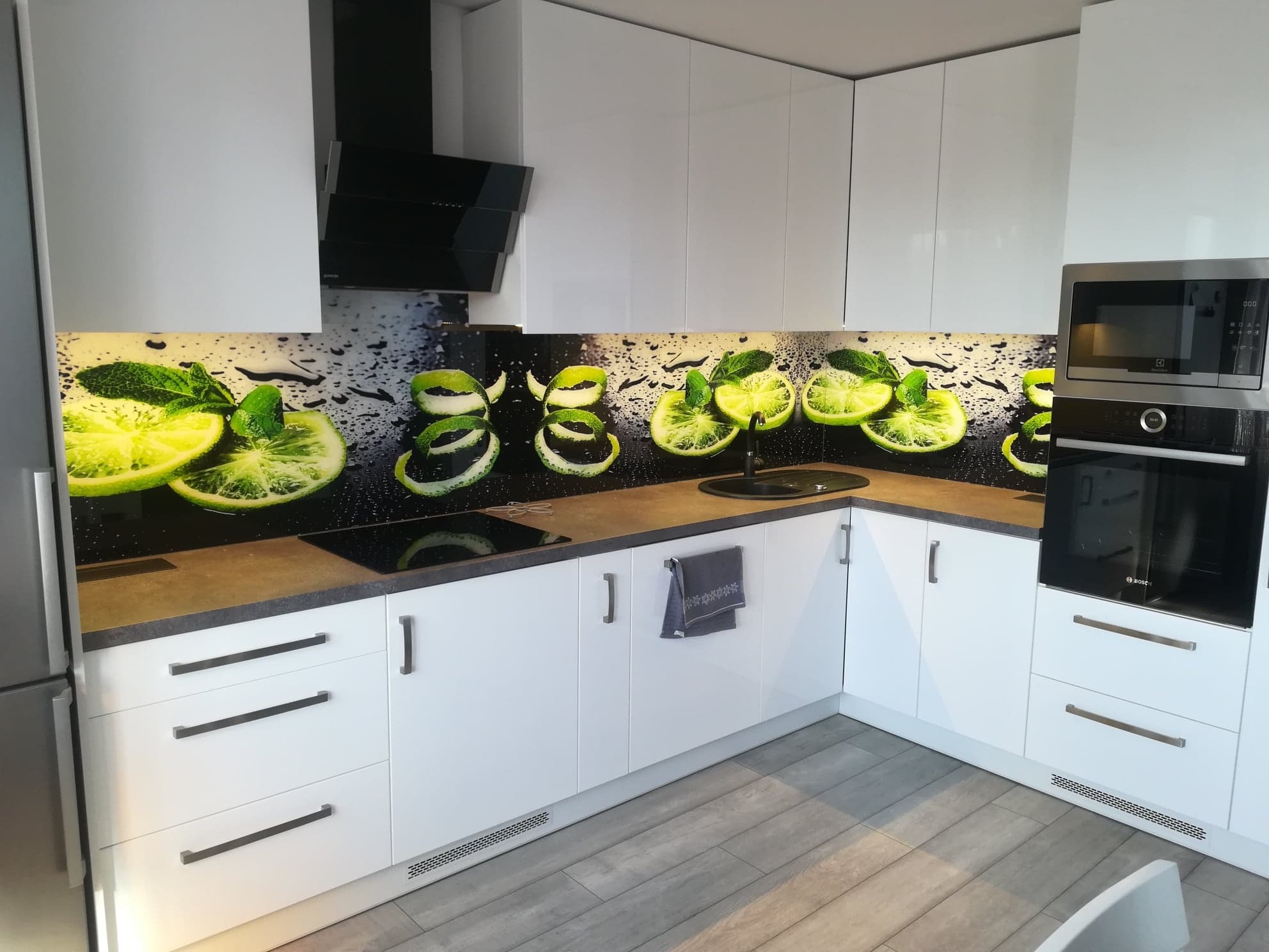 Biała kuchnia z błyszczącymi frontami i szarym blatem, ozdobiona panelem z motywem limonki i kropli deszczu, z widocznym piekarnikiem i płytą indukcyjną.
