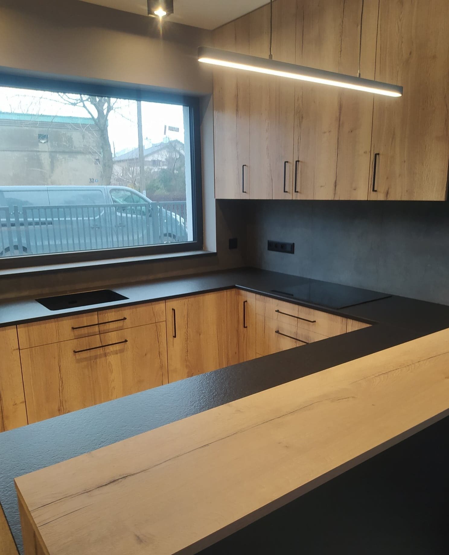 Nowoczesna kuchnia z drewnianymi frontami szafek i czarnym blatem, oświetlona podłużną lampą LED, z widokiem na ulicę przez okno.