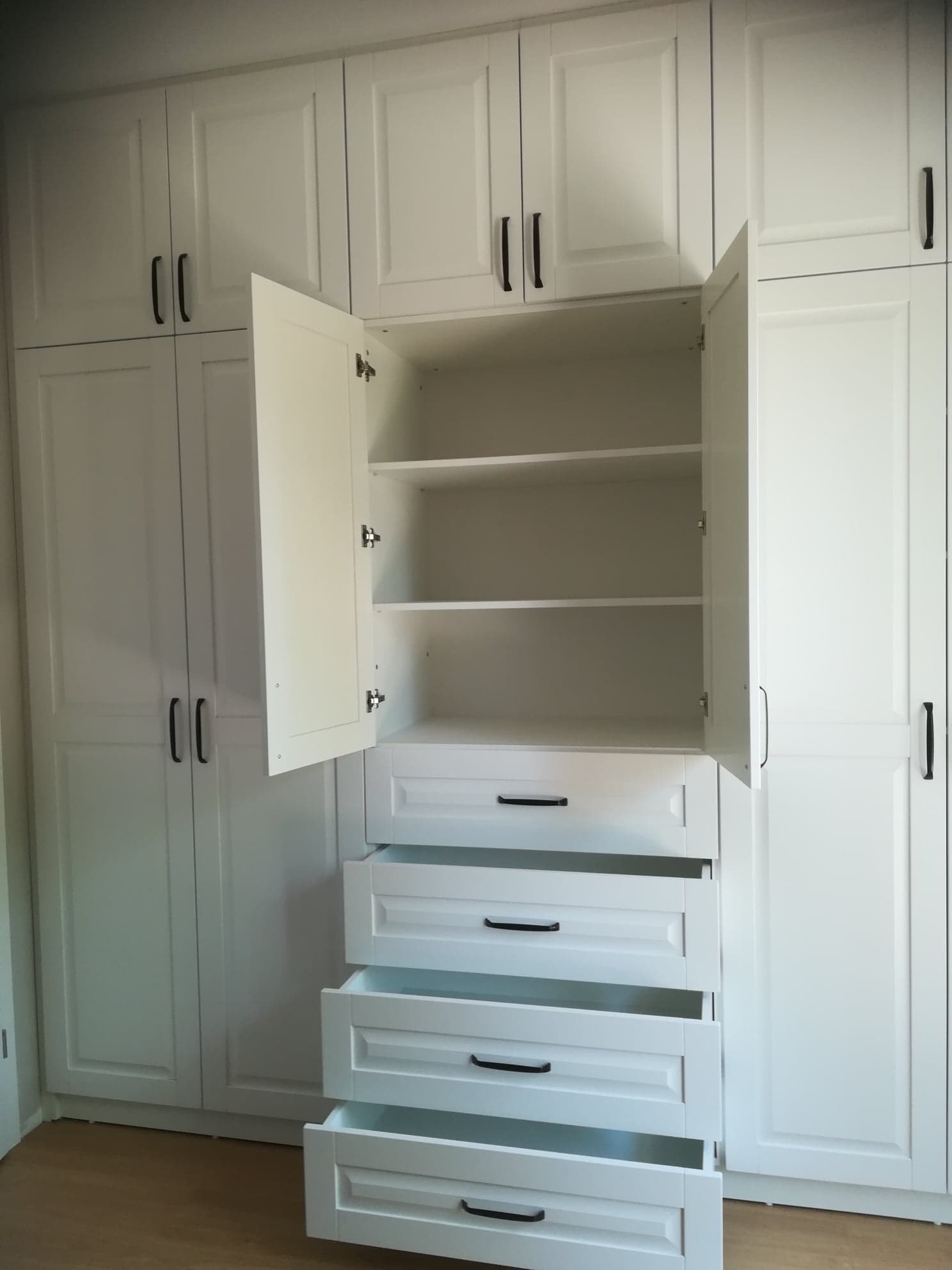 Biała zabudowa meblowa z szafkami i szufladami, otwarte drzwiczki ukazują puste półki, minimalistyczne czarne uchwyty.