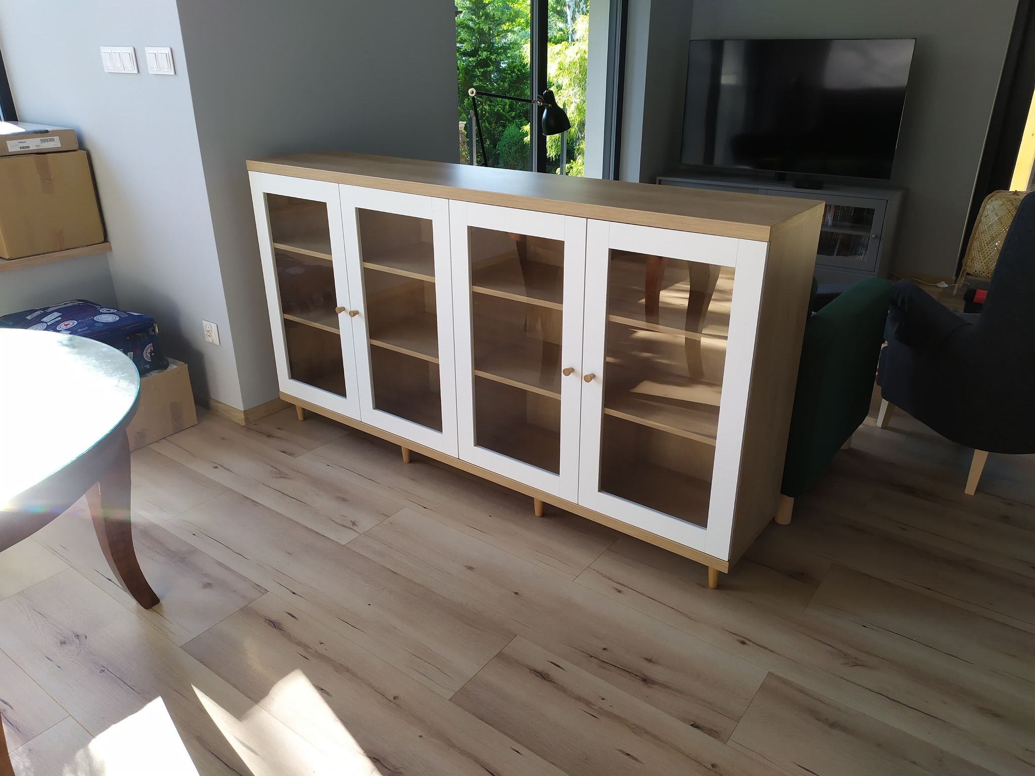 Nowoczesna przeszklona komoda z drewnianymi półkami i białymi ramami, ustawiona na niskich nóżkach w salonie z drewnianą podłogą.