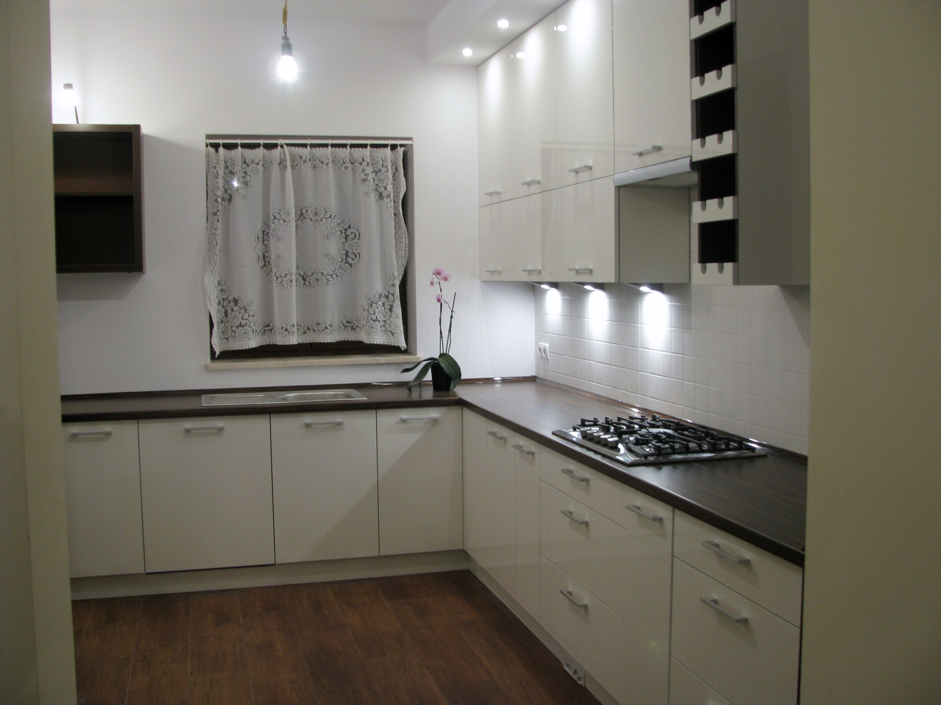 Jasna kuchnia z białymi, lakierowanymi szafkami z ciemnym blatem, widoczna płyta gazowa, okno z koronkową firanką i storczykiem, brązowa podłoga.