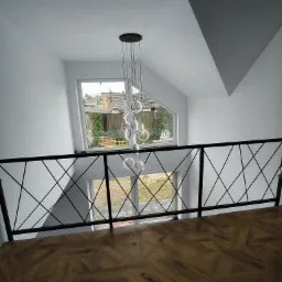 Wnętrze domu z drewnianą podłogą w jodełkę, czarną balustradą z geometrycznym wzorem i nowoczesną lampą wiszącą z okrągłych elementów, widok z góry na okno z widokiem na ogród.