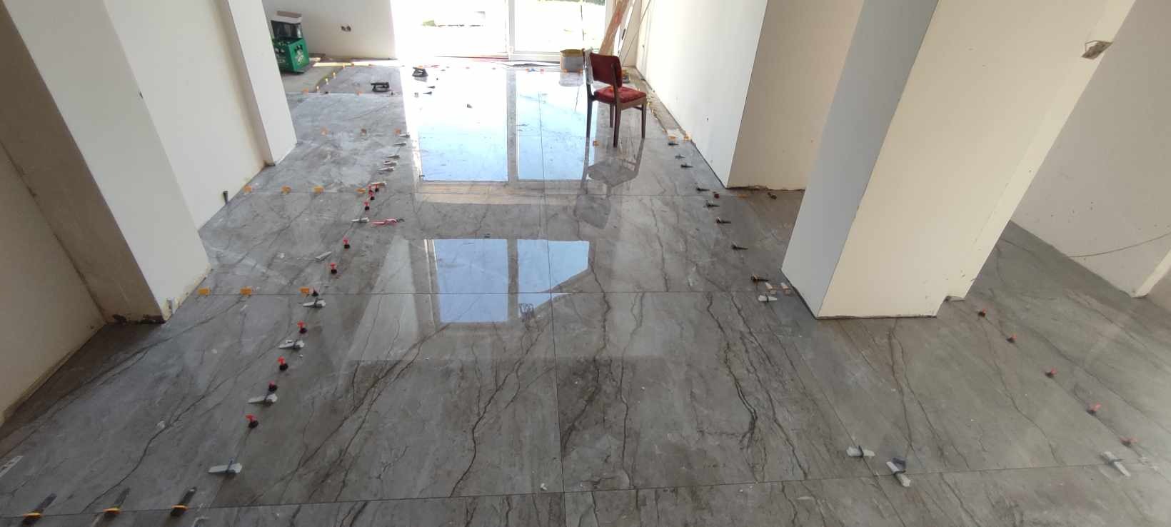 Układanie płytek podłogowych imitujących marmur w jasnym pomieszczeniu, widoczne plastikowe krzyżyki dystansowe i poziomica laserowa.