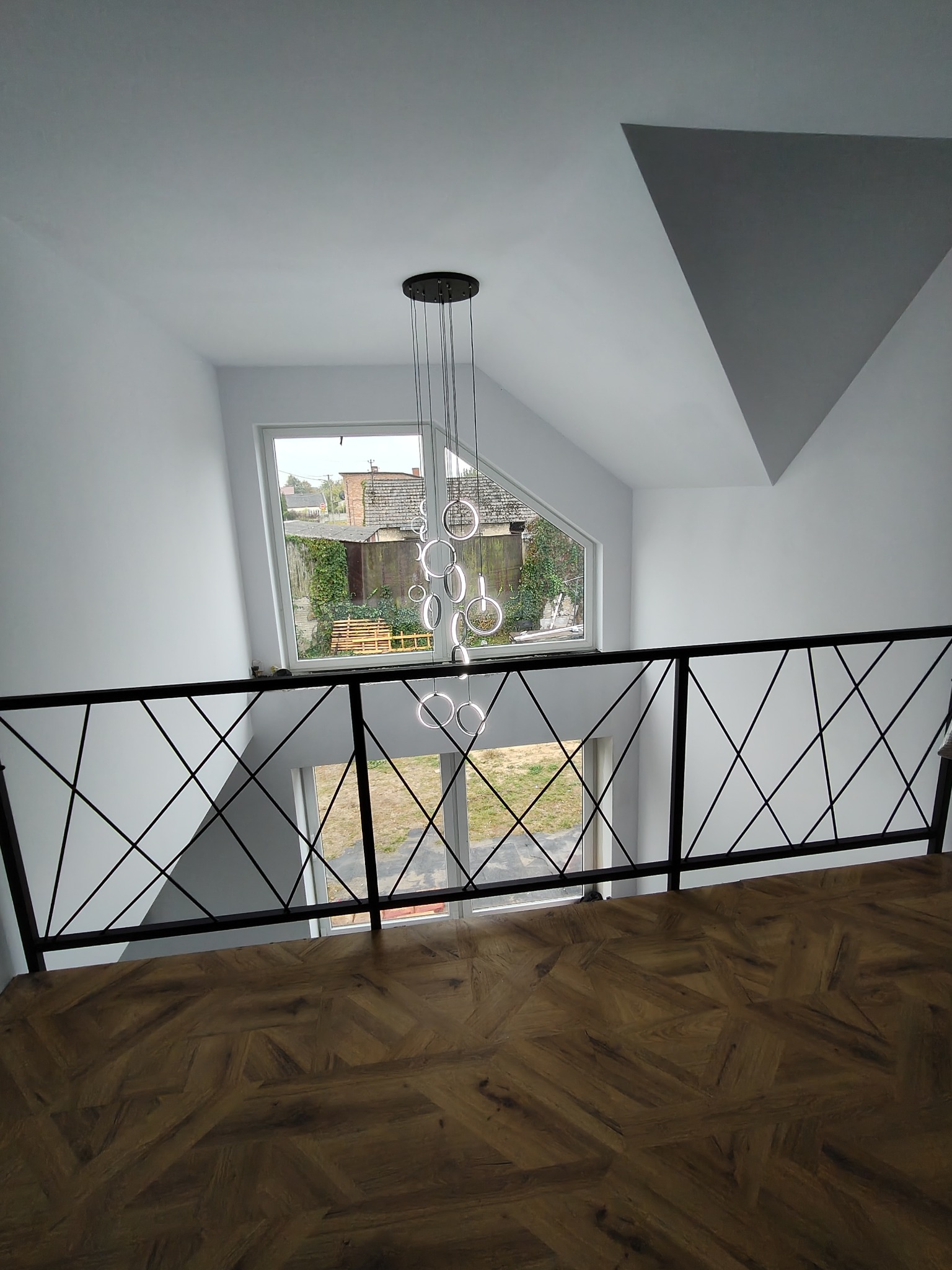 Wnętrze domu z drewnianą podłogą w jodełkę, czarną balustradą z geometrycznym wzorem i nowoczesną lampą wiszącą z okrągłych elementów, widok z góry na okno z widokiem na ogród.