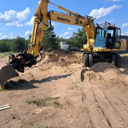 Żółta koparka kołowa Komatsu PW170ES z hydraulicznym chwytakiem, pracująca na piaszczystym terenie z widocznymi śladami opon i wykopem w tle, błękitne niebo z chmurami.