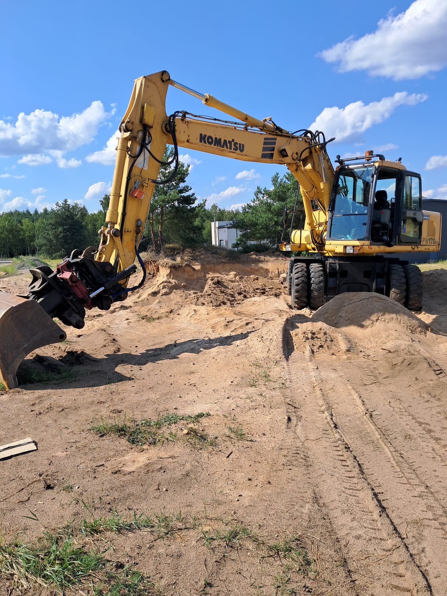 Żółta koparka kołowa Komatsu PW170ES z hydraulicznym chwytakiem, pracująca na piaszczystym terenie z widocznymi śladami opon i wykopem w tle, błękitne niebo z chmurami.