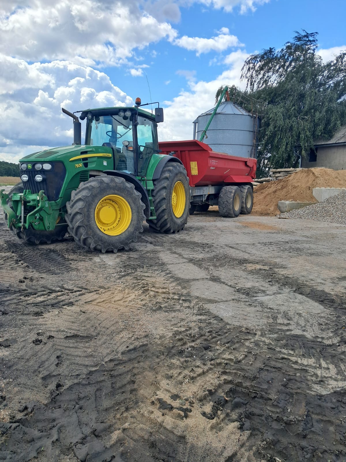 Zielony traktor John Deere 7930 z czerwoną przyczepą typu Kipper stoi na ubitym terenie obok kopca piasku i żwiru, w tle budynek gospodarczy i drzewo pod błękitnym niebem z chmurami.