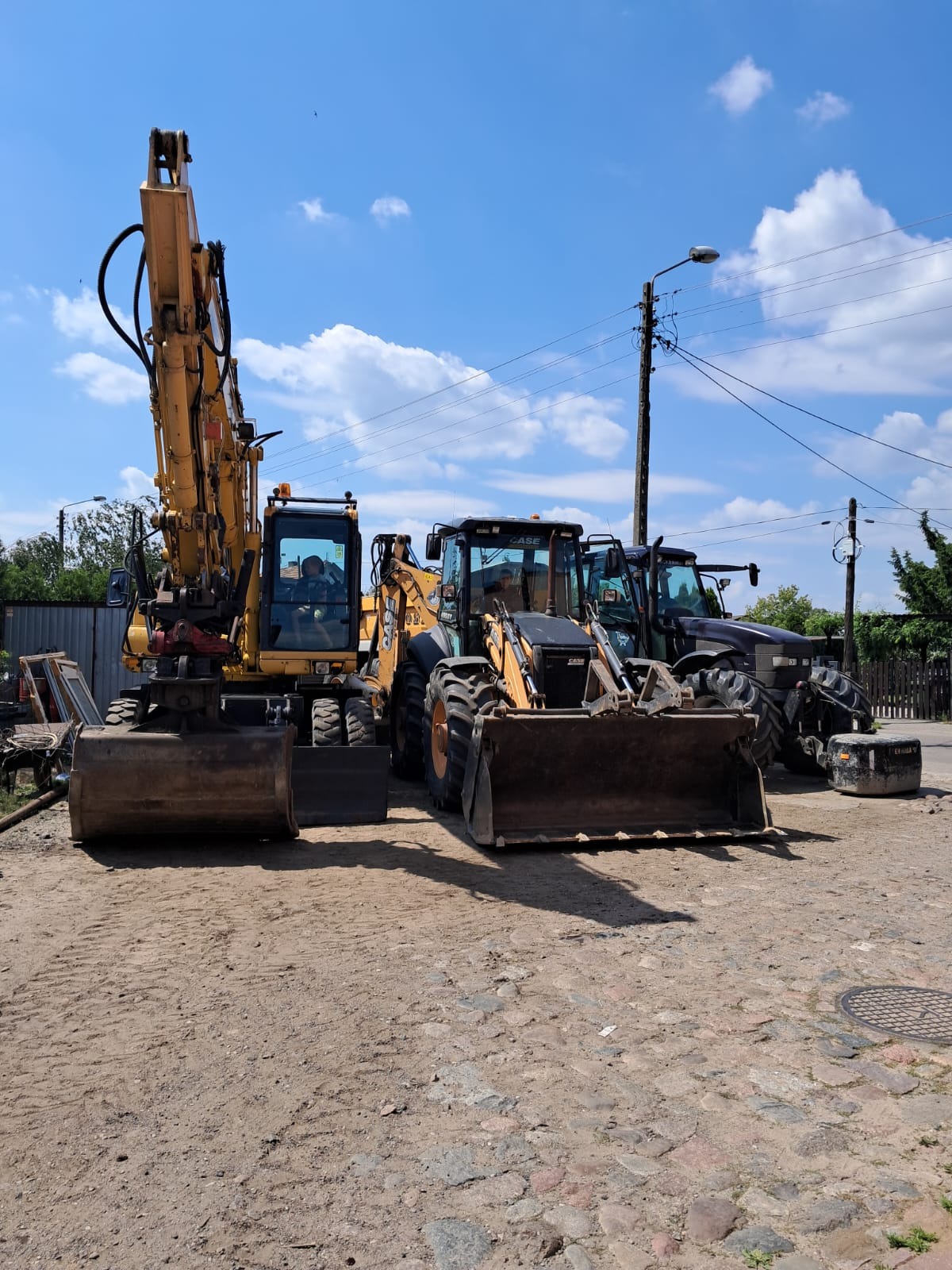 Żółty koparko-ładowarka i dwie koparki Case ustawione na placu budowy, widoczne ślady użytkowania i zabrudzenia na maszynach, w tle słup elektryczny i błękitne niebo z chmurami.