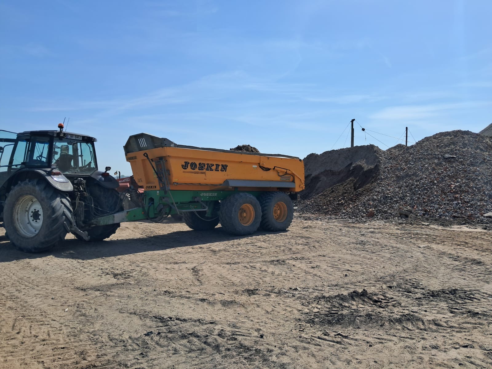 Żółta przyczepa Joskin załadowana ziemią, ciągnięta przez traktor na placu budowy, obok hałda gruzu i ziemi pod błękitnym niebem.