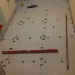 Układanie płytek podłogowych w łazience, widoczny poziomica, krzyżyki dystansowe i wiadro z wodą.