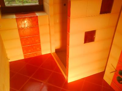 Wykończona łazienka z kabiną prysznicową, wyłożona płytkami w odcieniach czerwieni i beżu, z widocznym oknem i elementami dekoracyjnymi.